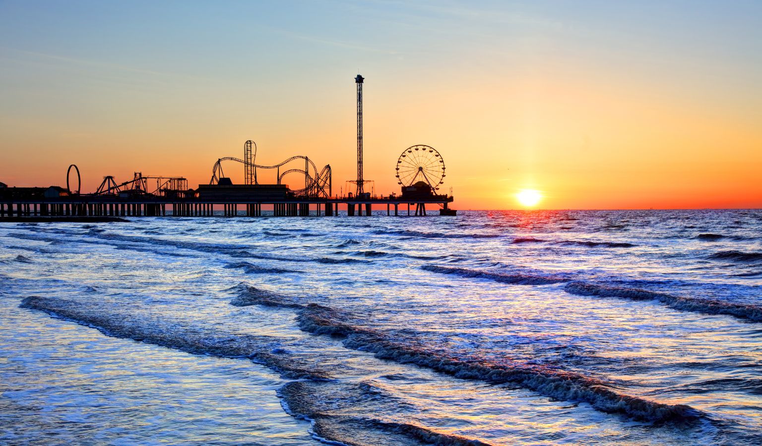 A vivid sunrise over Galveston Island, Texas, with gentle waves rolling toward the shore and the silhouette of the Galveston Island Historic Pleasure Pier — including its Ferris wheel and roller coaster — outlined against an orange and blue sky over the Gulf of Mexico.