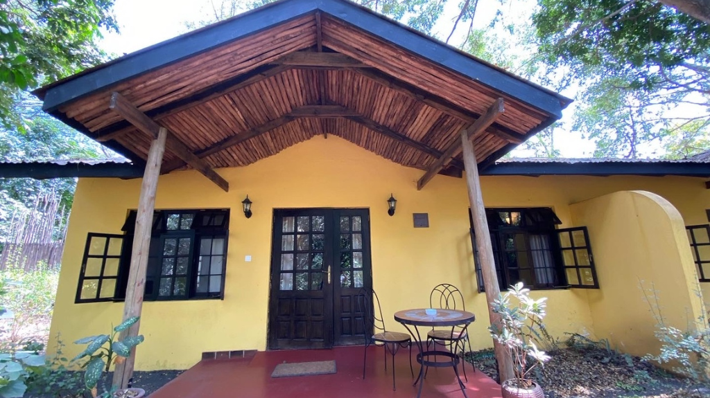 A charming yellow bungalow with a thatched roof and wooden supports, nestled in a lush, green area. The front has a small porch with a round table and chairs. The bungalow is accessible, where Laura and her friend stayed during their safari. Large windows provide a bright, airy feel to the accommodation.