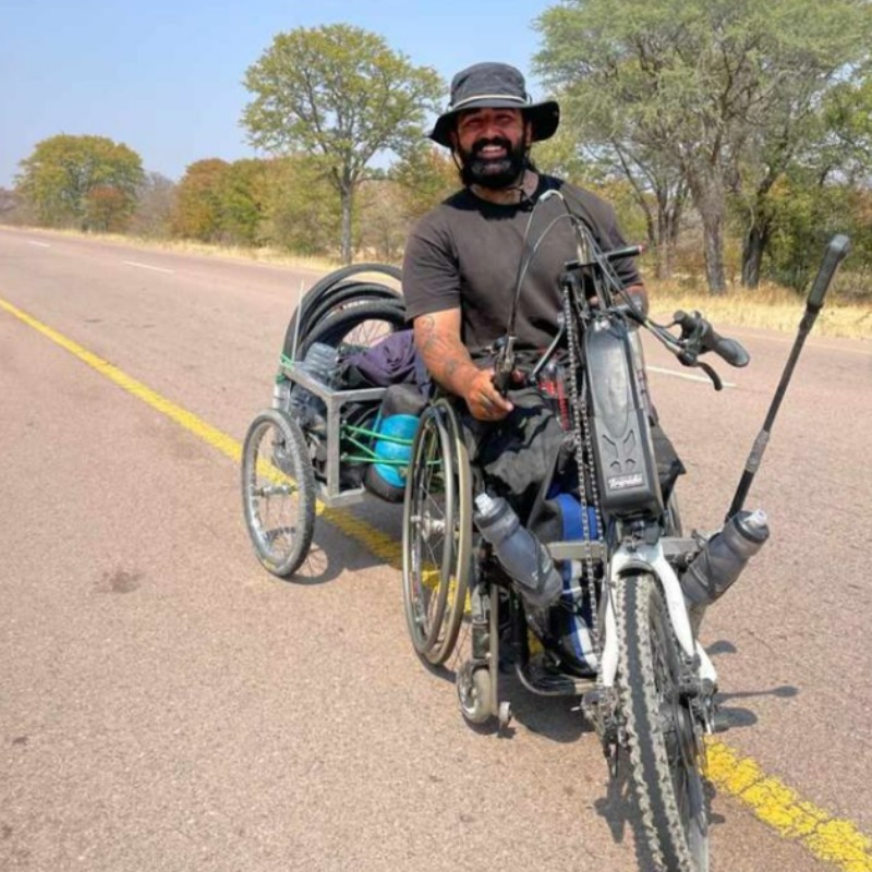 Schwan Wahab is seen smiling while on his journey, traveling along a remote road with sparse trees in the background. He is seated in his wheelchair, which has a handbike attachment on the front, and a homemade trailer cart at the back carrying his supplies. Schwan, dressed in a black shirt and wide-brimmed hat, appears ready for the long journey ahead. The image captures the spirit of his monumental challenge as he navigates African travel for wheelchair users, demonstrating both resilience and ingenuity on his path across the continent.