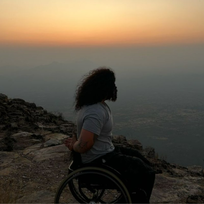 Schwan Wahab sits in his wheelchair on a rocky cliffside, gazing out at a hazy sunset over distant mountains and a vast landscape. His back is to the camera, and his long, curly hair flows in the breeze. The serene setting highlights the peaceful yet adventurous nature of his journey as he continues his wheelchair travel adventures across the African continent. The image captures a moment of reflection, embodying the spirit of exploration and determination that defines his travels.