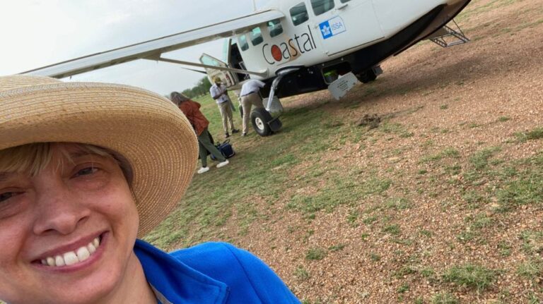 Laura smiles in the foreground, wearing a wide-brimmed hat and blue jacket, standing in front of a small Coastal aviation Cessna plane on a dirt airstrip during a safari in Tanzania. Although the plane isn't the most accessible, Laura was able to manage boarding with some assistance.