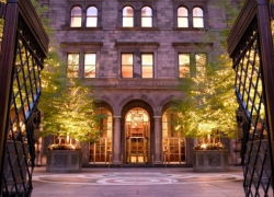 An elegant entrance of the accessible Lotte New York Palace Hotel in Manhattan, framed by wrought iron gates. The historic facade features large arched windows, with warm lighting illuminating the building and surrounding trees, creating a welcoming atmosphere.