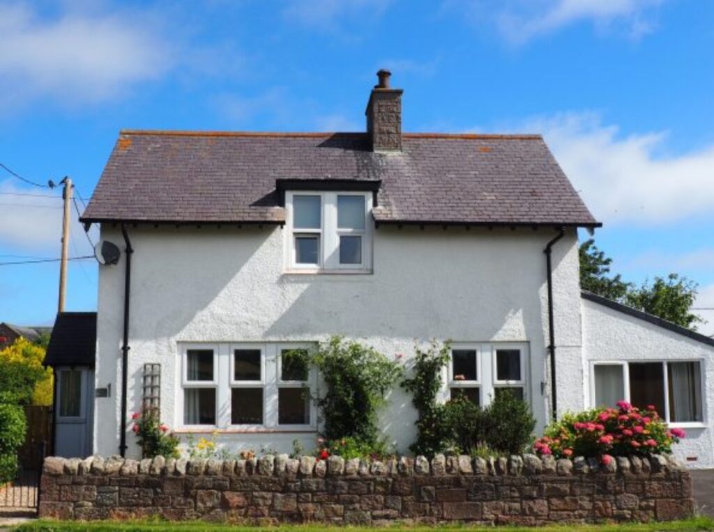 [Image Description: A charming, traditional white two-story house with a dark, sloping roof and a central chimney. It has a row of four windows on the ground floor, with the two center ones slightly wider, and a pair of windows on the upper floor. The house is surrounded by a low stone wall, with flowers and greenery decorating the front garden, adding a touch of color against the clean white facade.]