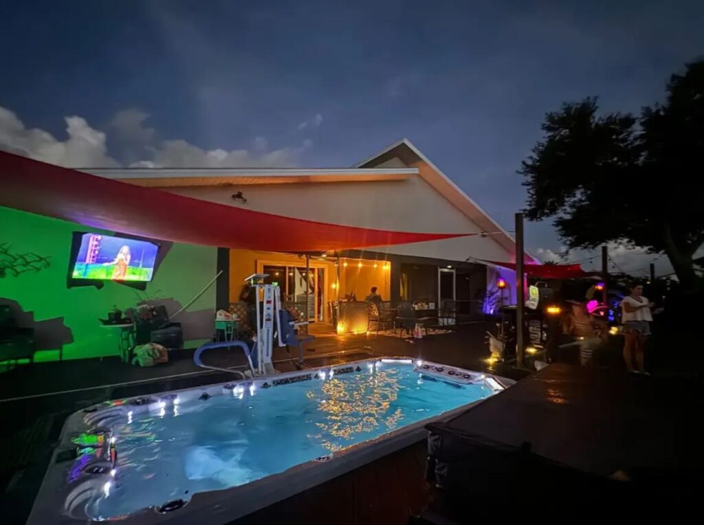 Evening view of an accessible villa's backyard in Cape Coral, featuring a well-lit swim spa with a pool lift and a mounted outdoor TV. The patio area has comfortable seating, string lights, and red sun shades, creating a cozy and inviting atmosphere.