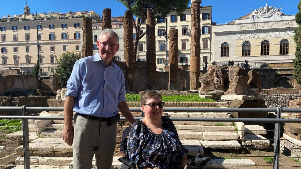 A man and a woman are posing in front of ancient Roman ruins on a sunny day. The woman, seated in a wheelchair, smiles, while the man stands beside her, also smiling. Behind them are tall, worn columns and stone structures, with a historic building in the background. The scene conveys an accessible historical site.