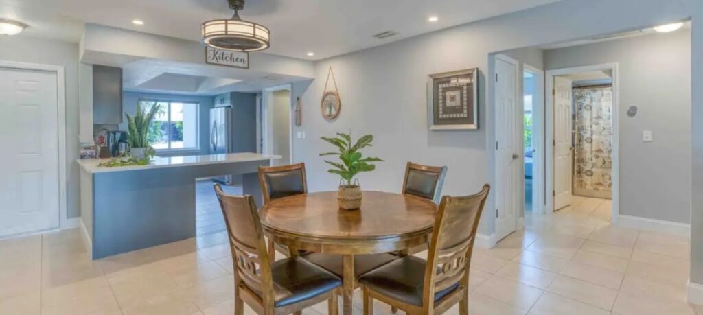 Bright and open dining area in an accessible Cape Coral villa, featuring a round wooden table with four chairs, a decorative plant centerpiece, and a view into the modern kitchen with gray cabinets. The space has light-colored tile flooring and tasteful decor on the walls, creating a welcoming ambiance.