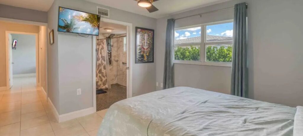 Bedroom in the accessible Cape Coral villa with a comfortable bed, wall-mounted TV, and large window with curtains offering natural light. An open doorway leads to a spacious bathroom with a roll-in shower, designed with accessibility in mind. The room has a calm, welcoming atmosphere and is well-suited for relaxation.