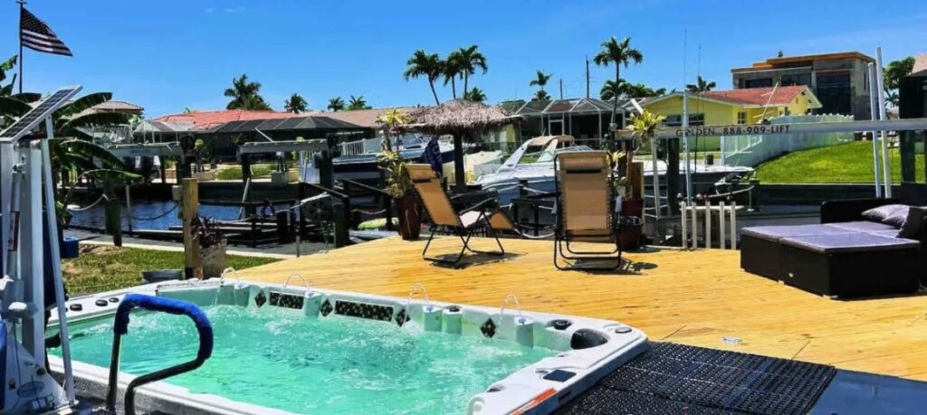 Outdoor deck area at the accessible Cape Coral villa, featuring a swim spa with a pool lift, lounge chairs, and a view of the canal with boats docked nearby. The deck is surrounded by lush tropical plants and has easy access to the water, creating a relaxing and inviting space for sunbathing and enjoying the Florida sunshine.