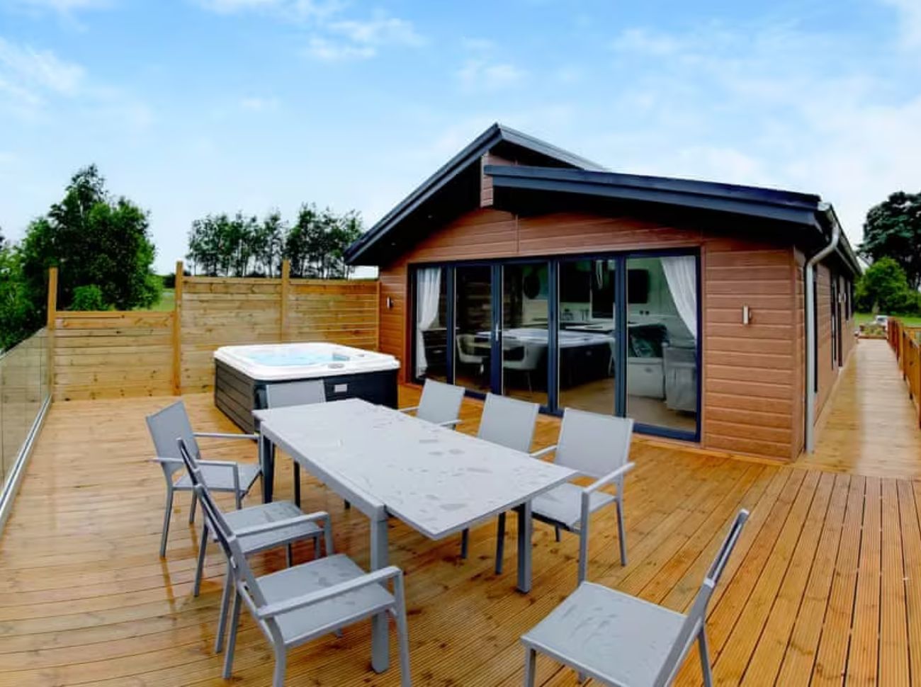 A wheelchair-friendly cabin in Lincolnshire with a spacious wooden deck featuring outdoor seating for six and a private hot tub. The accessible holiday home is surrounded by greenery and large windows offering a view of the interior amenities.