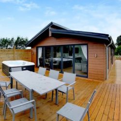 A modern wheelchair-friendly cabin at Noah's Retreat in Lincolnshire featuring a spacious wooden deck with outdoor seating for six and a private hot tub. This accessible holiday home offers a relaxing retreat surrounded by greenery.