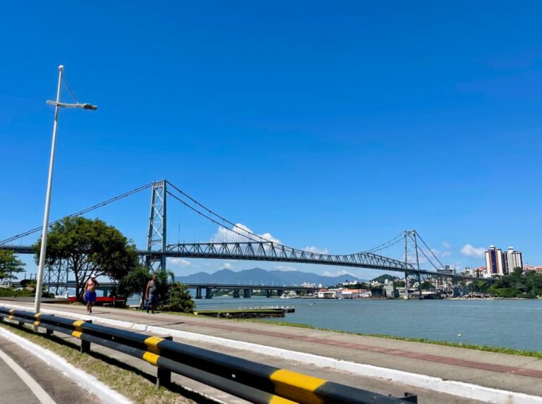 A bright, sunny day with a clear blue sky showcasing a suspension bridge spanning across a calm river. The foreground features a pedestrian walkway with a black and yellow guardrail, bordered by a grassy area with trees. In the background, buildings and a mountainous landscape can be seen.