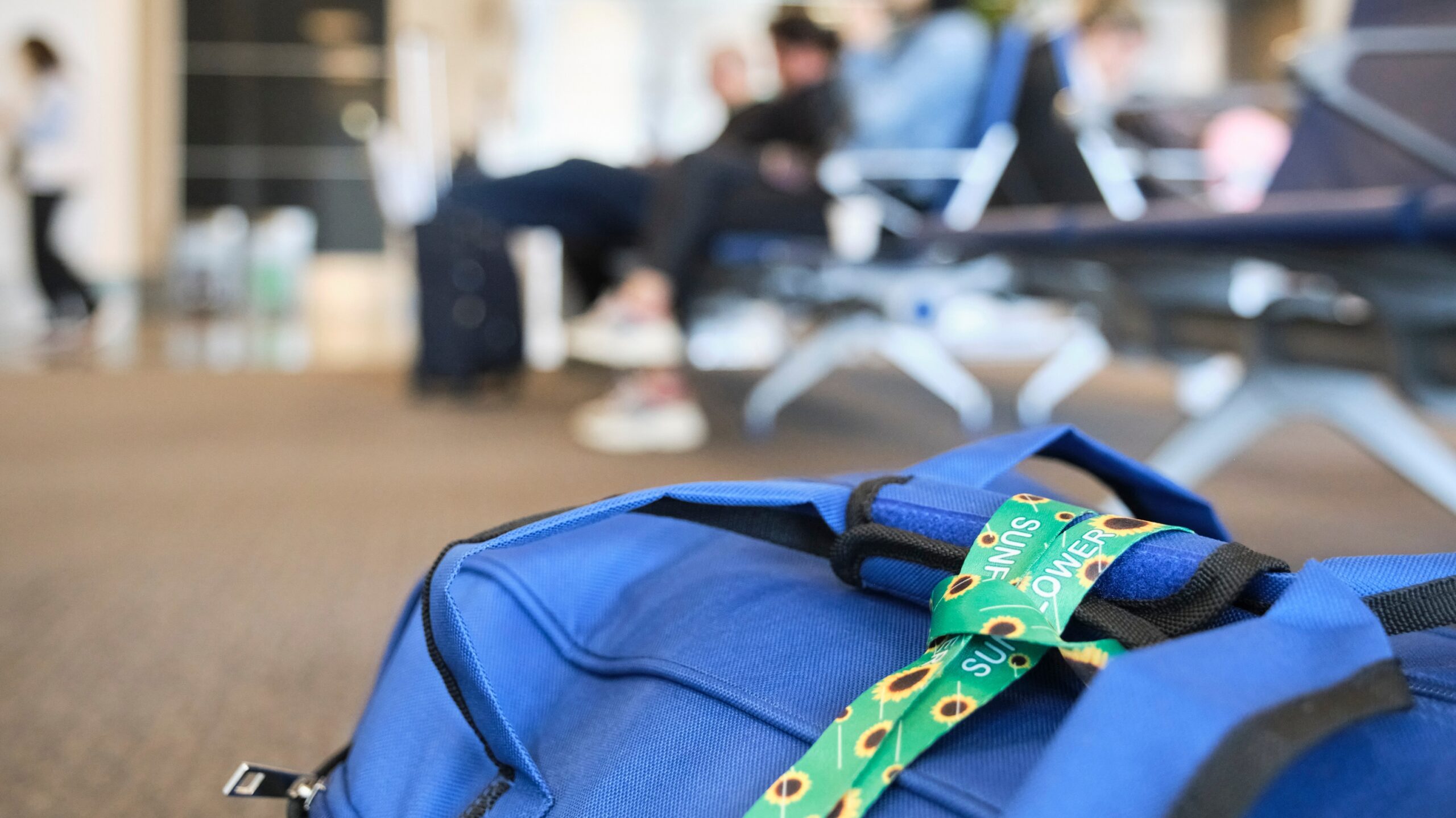 A close-up of a bright blue bag sitting on the floor in an airport lounge. Attached to the bag is a green lanyard with yellow sunflower patterns, commonly associated with hidden disabilities. In the blurred background, travelers are seated on benches, creating a busy yet calm airport atmosphere.