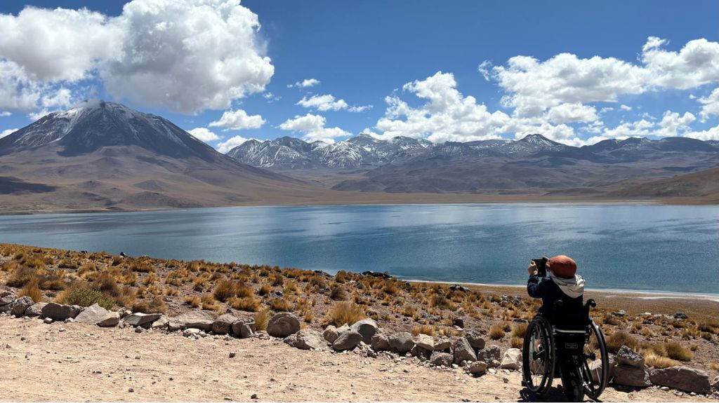 Joan Pahisa sits in his wheelchair with his back to camera, taking a photograph of Laguna Miscanti.