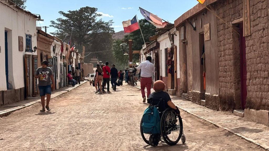 Joan sits in his chair, admiring the streets of San Pedro de Atacama. The streets are very dusty with small buildings lining on either side. The flag of Chile hangs from several of the buildings.