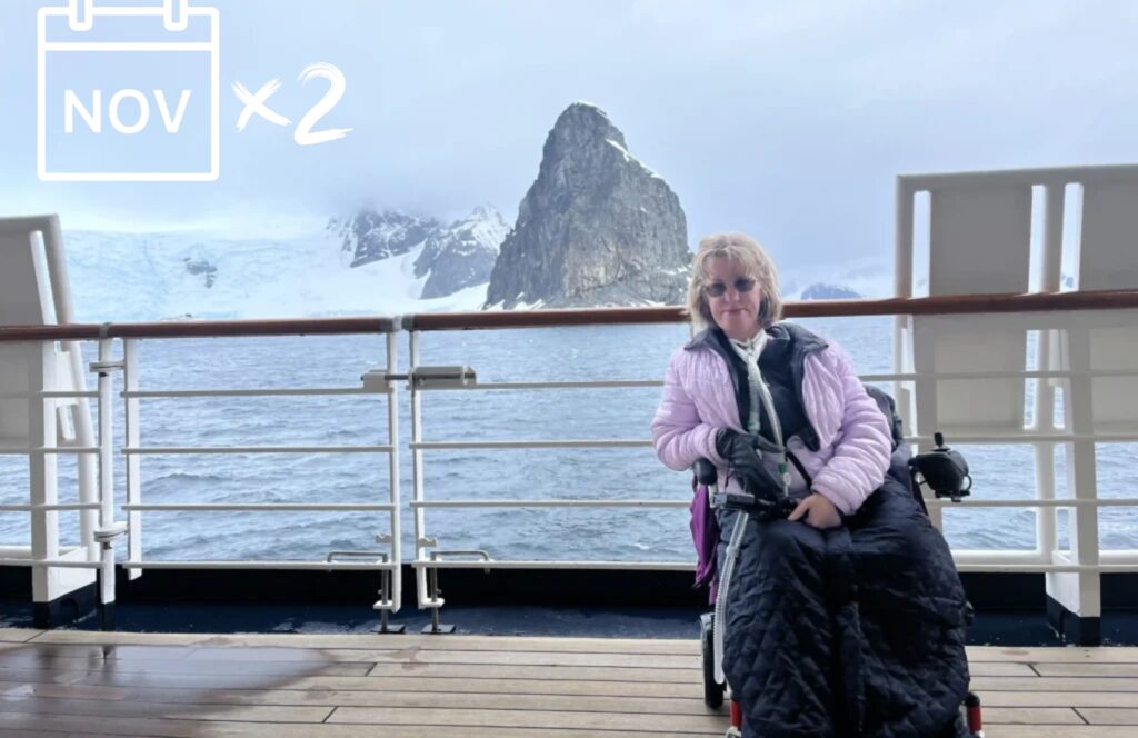 A woman in a power wheelchair, bundled in a black wheelchair blanket and wearing a lavender jacket, smiles while on the deck of a cruise ship with dramatic snow-covered mountains and icy waters in the background. A calendar icon marked "NOV x2" appears in the top left, possibly referencing two November events or editions. The scene captures the spirit of accessible adventure in cold, remote destinations.