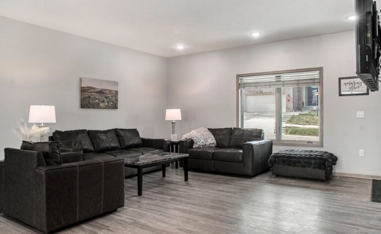 Large open living room with grey wood-style flooring, featuring a dark leather sectional sofa, marble-topped coffee table, and bright recessed lighting.