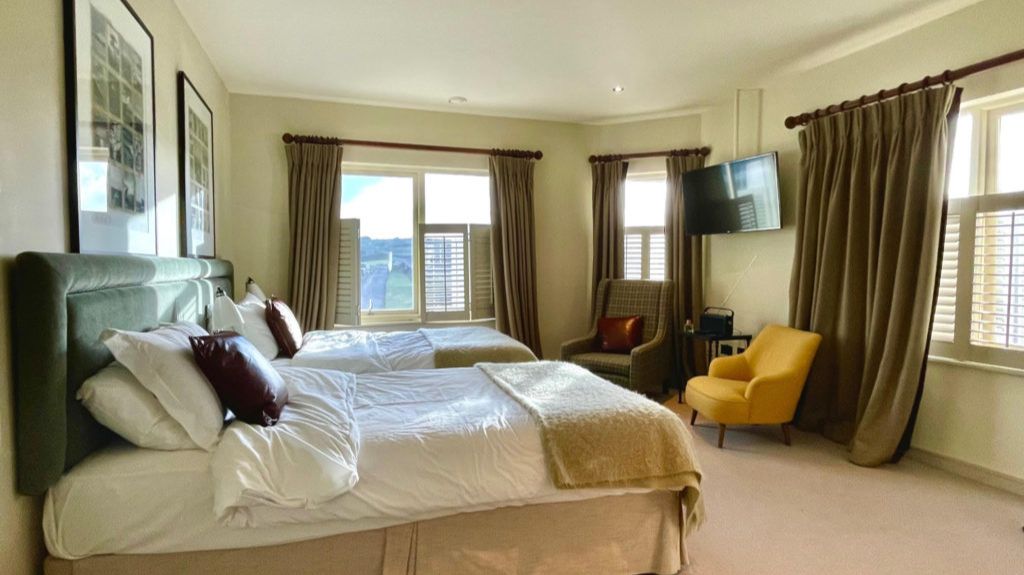 A spacious, well-lit hotel room with two double beds featuring white linens and brown accent pillows. The room has large windows with beige curtains, a wall-mounted TV, a gray armchair with a brown cushion, and a mustard-yellow armchair. Framed artwork hangs on the wall above the beds, and the decor is modern and neutral-toned.