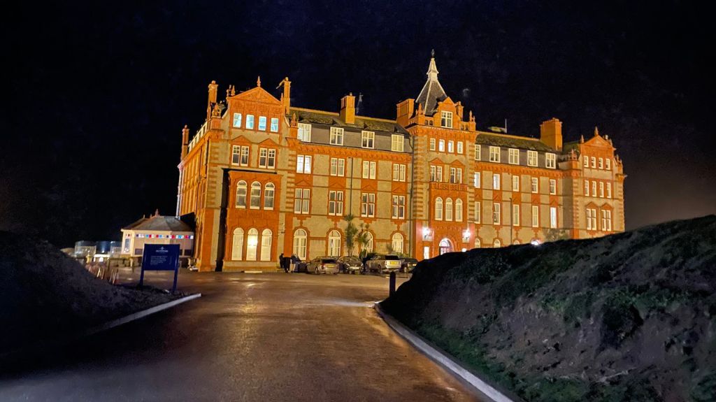 A large, historic red-brick building illuminated at night. The multi-story structure features ornate architectural details, arched windows, and a central tower with a pointed roof. The building is set against a dark sky, with a paved driveway leading toward the entrance. A few cars are parked in front, and a small building with string lights is visible to the side.