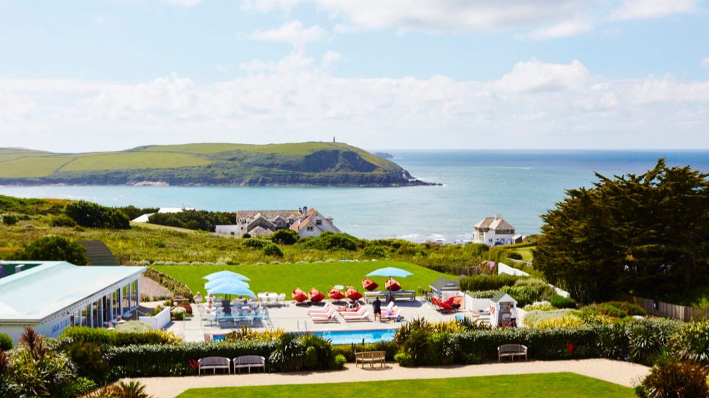 A scenic coastal view featuring a hotel or resort pool area with red sun loungers and blue umbrellas, surrounded by manicured lawns and lush greenery. In the background, the ocean stretches out under a partly cloudy sky, with a green headland jutting into the sea. Several white houses are dotted along the coastline.