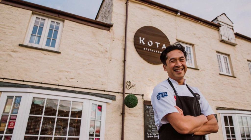 A smiling chef stands with arms crossed in front of Kota Restaurant, housed in a rustic cream-colored stone building with white-framed windows. The restaurant's round wooden sign is visible above the chef. The chef wears a white shirt with branded patches and a dark apron.