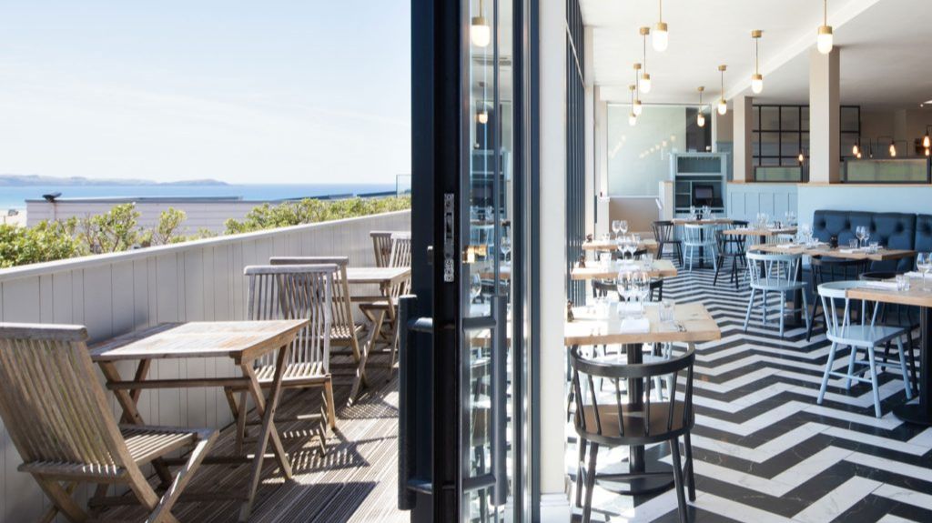 A bright, modern restaurant with indoor and outdoor seating. The interior features black and white chevron-patterned flooring, light wooden tables, and a mix of black and light blue chairs. Pendant lights hang from the ceiling. Outside, wooden tables and chairs are set on a deck overlooking greenery and the ocean in the distance under a clear sky.