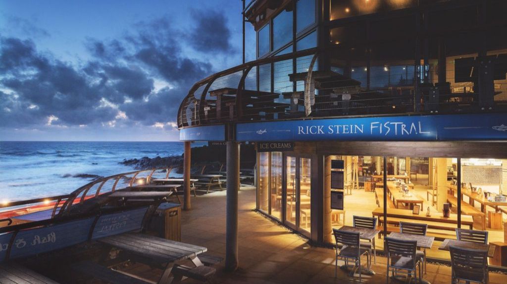 A seaside restaurant called Rick Stein Fistral, with large glass windows revealing a warmly lit interior filled with wooden tables and chairs. The exterior features a balcony with additional seating, overlooking the ocean. The scene is at dusk, with dramatic clouds in the sky and waves crashing against the rocky shoreline.