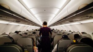 A flight attendant walks down the aisle of a full airplane cabin, seen from behind, with passengers seated on both sides.