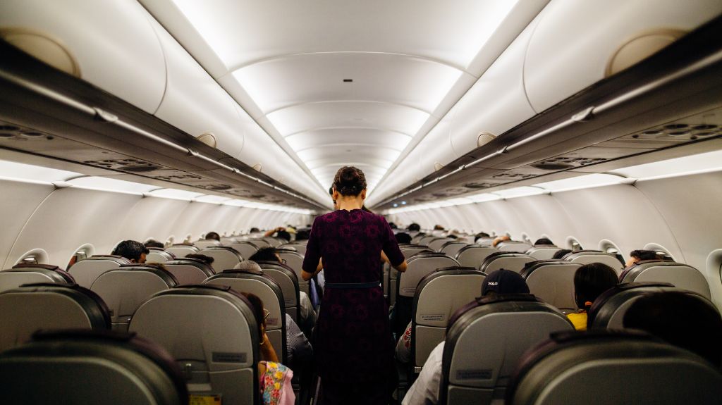 A flight attendant walks down the aisle of a full airplane cabin, seen from behind, with passengers seated on both sides.