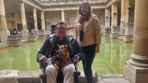 Man using a wheelchair with a small dog on his lap and a woman standing beside him, smiling in front of the Roman Baths in Bath, England.