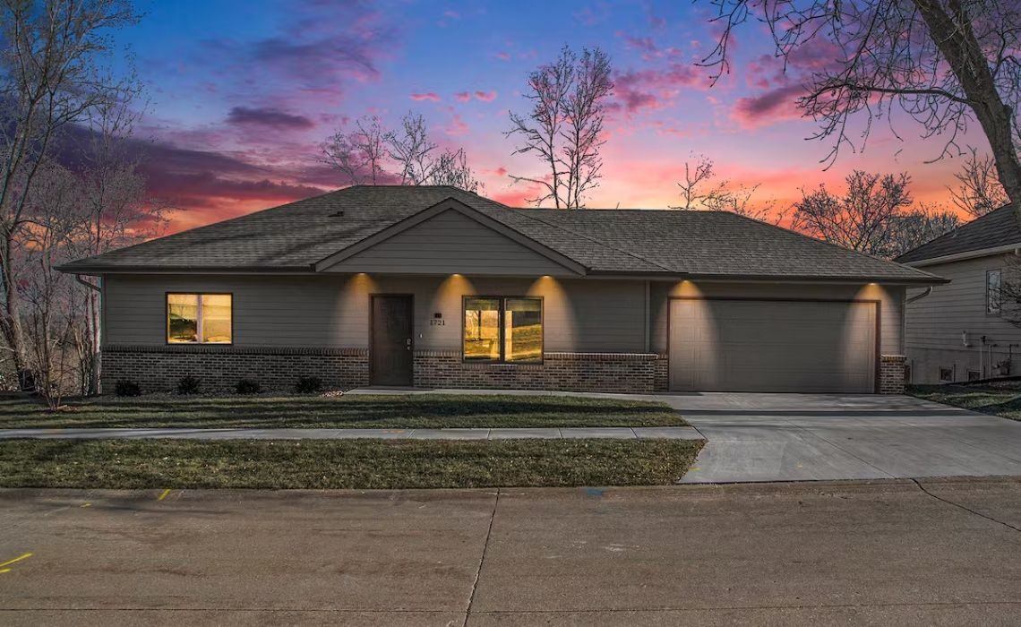 Front exterior of the Lemonade vacation rental at sunset, featuring a modern ranch-style house with gray siding, brick accents, warm outdoor lighting, and a spacious two-car garage.