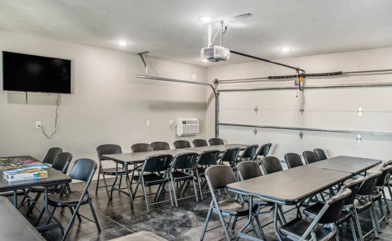 Versatile indoor space set up with multiple long folding tables and black chairs, perfect for group meetings or activities, equipped with a wall-mounted TV and air conditioning.