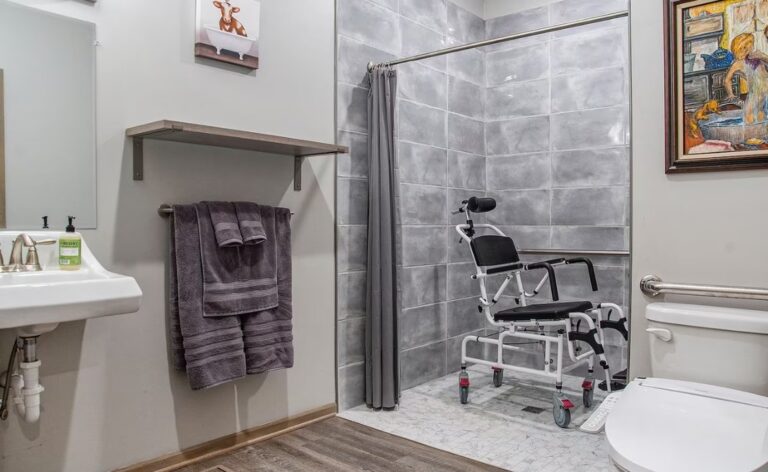 Accessible bathroom featuring a roll-in shower with grey tiling, a specialized white shower wheelchair with a headrest, grab bars by the toilet, and a wall-mounted sink for easy access.
