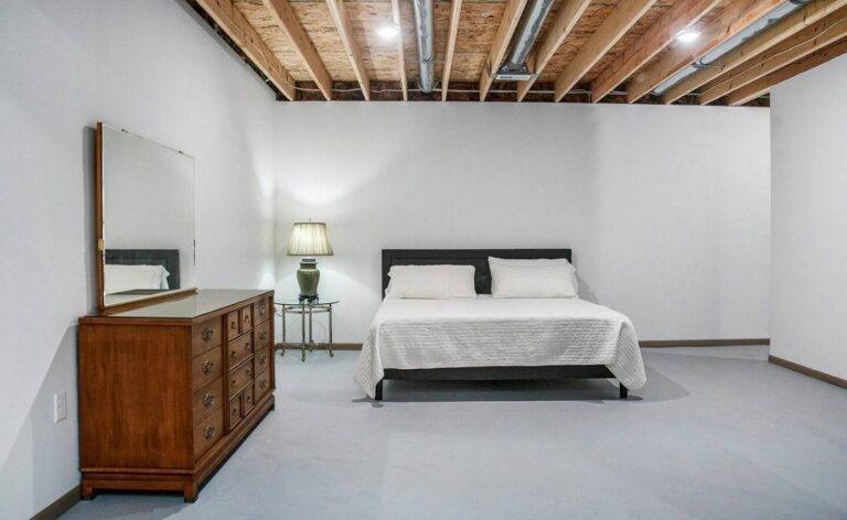 Spacious bedroom area in a finished basement with exposed ceiling beams, featuring a dark wood bed frame with white linens and a matching vintage wooden dresser with a large mirror.