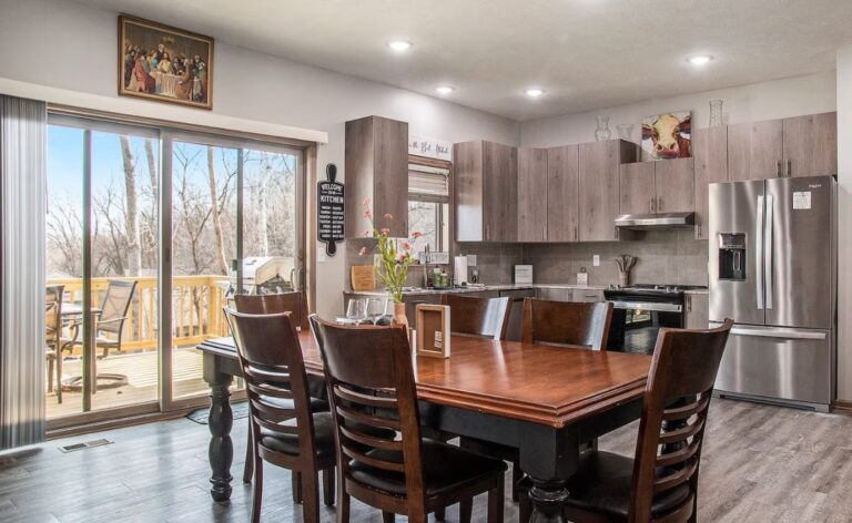 Open-concept dining area with a large dark wood table and six chairs, leading into a modern kitchen with grey cabinetry and stainless steel appliances.