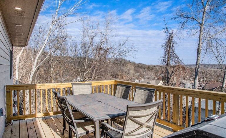 Wooden outdoor deck with a large dining table and six chairs, overlooking a scenic view of bare trees and a valley under a bright blue sky.