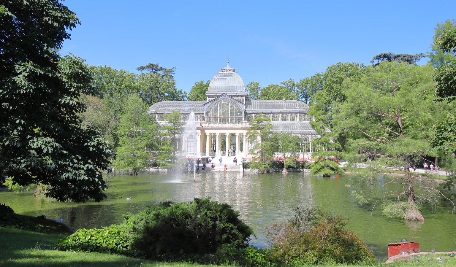 Crystal Palace in El Retiro Park, Madrid, viewed across the pond with fountain and surrounding trees.