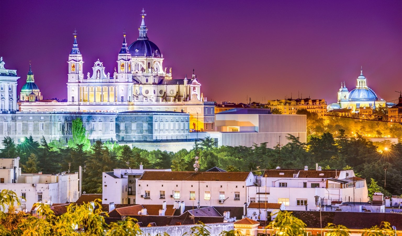 Night view of Almudena Cathedral in Madrid illuminated against a purple sky with city rooftops in the foreground.