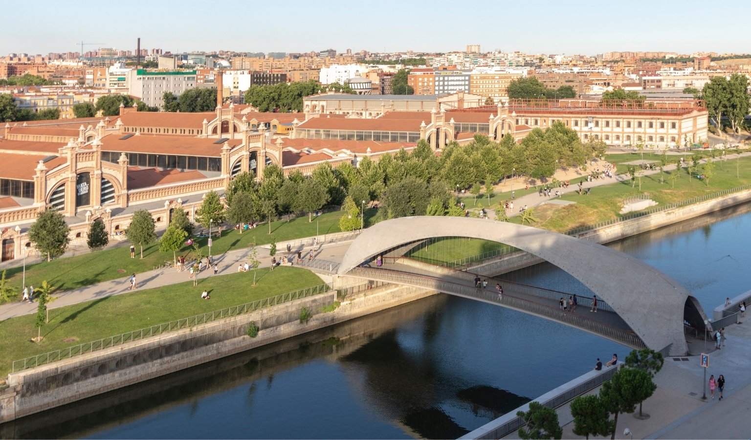 Modern pedestrian bridge crossing the Manzanares River at Madrid Río park with the historic Matadero cultural complex in the background.