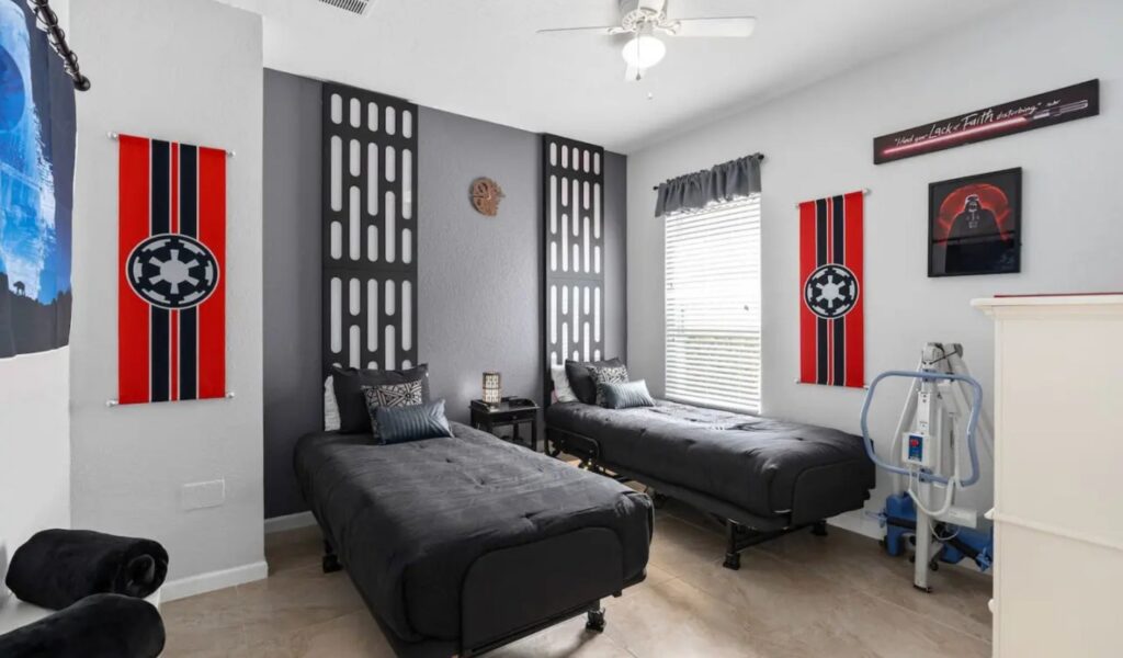 A Star Wars-themed bedroom with two twin beds featuring black bedding. The room is decorated with red Imperial banners, a Darth Vader framed poster, and custom wall panels reminiscent of a Death Star corridor. A mobile patient lift is positioned in the corner, and a lightsaber-themed wall plaque hangs above the bed.