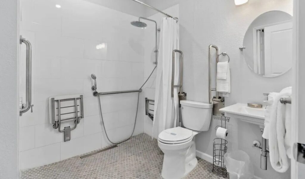 A modern, white-tiled bathroom designed for accessibility. It features a roll-in shower with a rainfall showerhead, a handheld sprayer, and fold-down wooden benches. Multiple stainless steel grab bars are installed around the shower and the toilet. A pedestal sink with a round mirror and a wall-mounted towel rack are also visible.