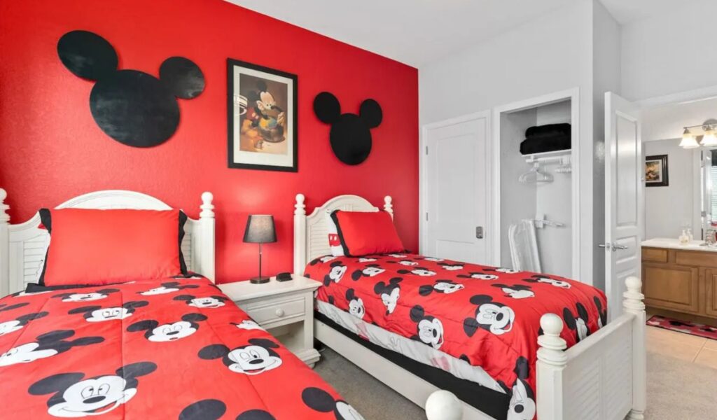 A bright bedroom featuring two twin beds with red Mickey Mouse patterned comforters. The focal wall is painted vibrant red and decorated with two large black Mickey Mouse silhouette cutouts flanking a framed Mickey Mouse vintage-style poster. Between the white headboards sits a small white nightstand with a black lamp.