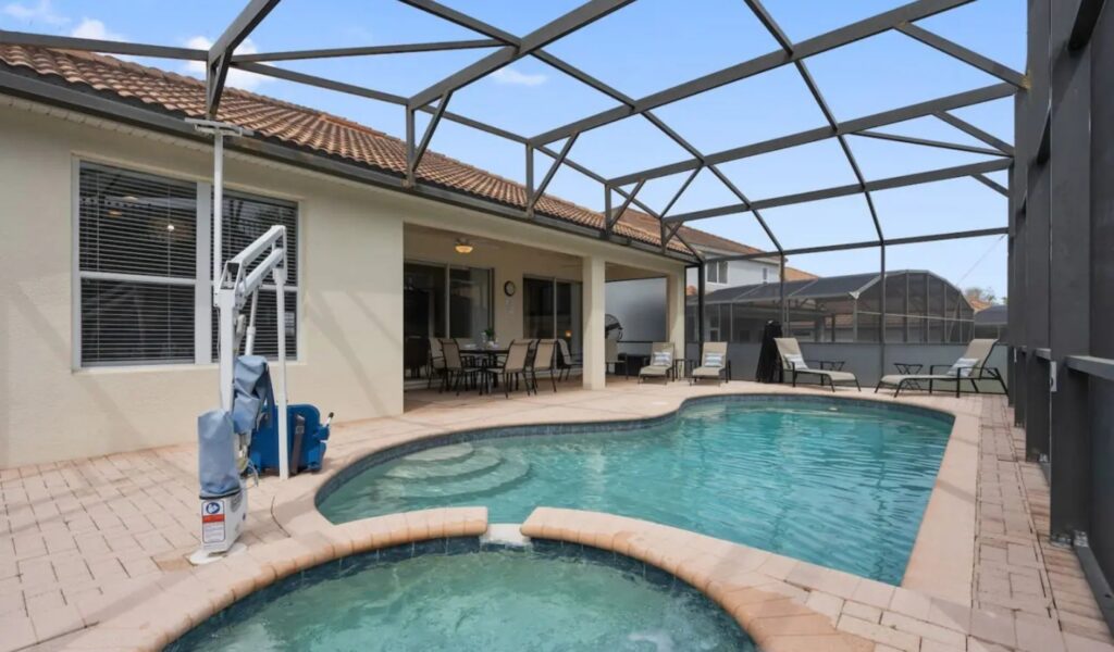 : A wide shot of a private, screened-in swimming pool and integrated spa. A white and blue motorized ADA pool lift is installed on the paved deck for accessible entry. The area includes several lounge chairs and a covered patio with an outdoor dining set under a beige exterior wall.