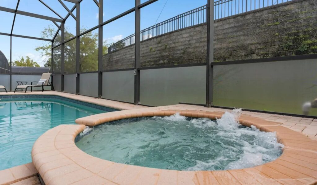 A close-up view of a circular, bubbling outdoor spa integrated into a larger swimming pool. The spa is surrounded by tan stone coping and sits within a screened-in lanai. A tall privacy wall with a metal fence is visible in the background against a clear blue sky.