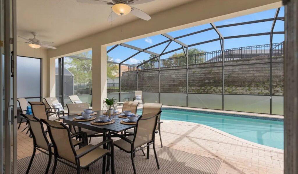 A shaded patio dining area featuring a dark rectangular table set for six with blue and white dishware. The space is cooled by ceiling fans and opens directly onto the sun-drenched pool deck and screened-in enclosure. A neutral-toned rug sits under the table, and additional lounge seating is visible in the background.