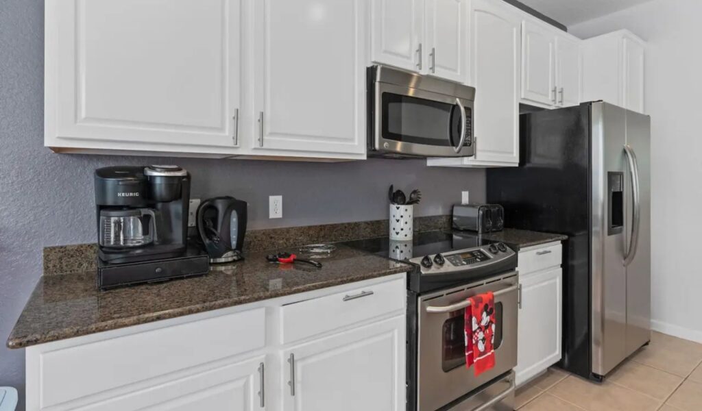 A close-up of a kitchen featuring white cabinetry and dark granite countertops. The space is equipped with stainless steel appliances, including a glass-top stove, over-the-range microwave, and a large side-by-side refrigerator. A Keurig coffee maker and electric kettle sit on the counter, and a red Mickey Mouse-themed kitchen towel hangs from the oven handle.