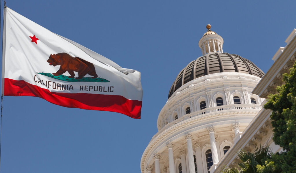 California state flag flying beside the white dome of the State Capitol building in Sacramento on a sunny day