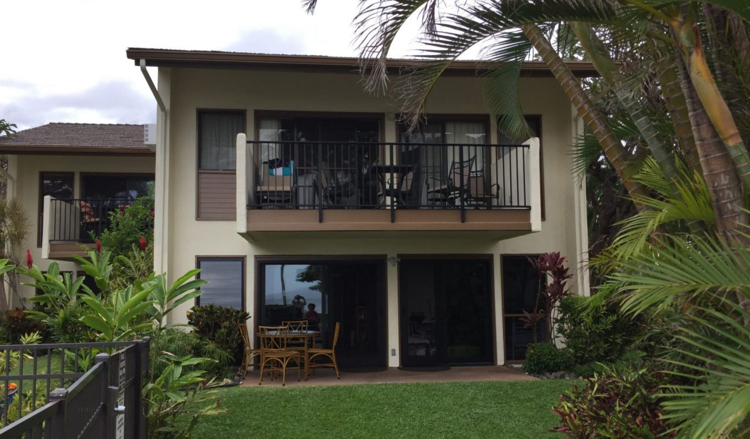 Exterior view of a ground-floor accessible condo in Maui, Hawaii, showing a well-maintained lawn, patio dining set, and wide sliding glass doors suitable for wheelchair access. Surrounded by lush tropical plants and palm trees.