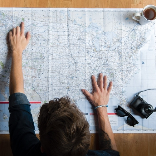 Traveler studying a large map of the United States with a camera, sunglasses, and cup of tea on the table.