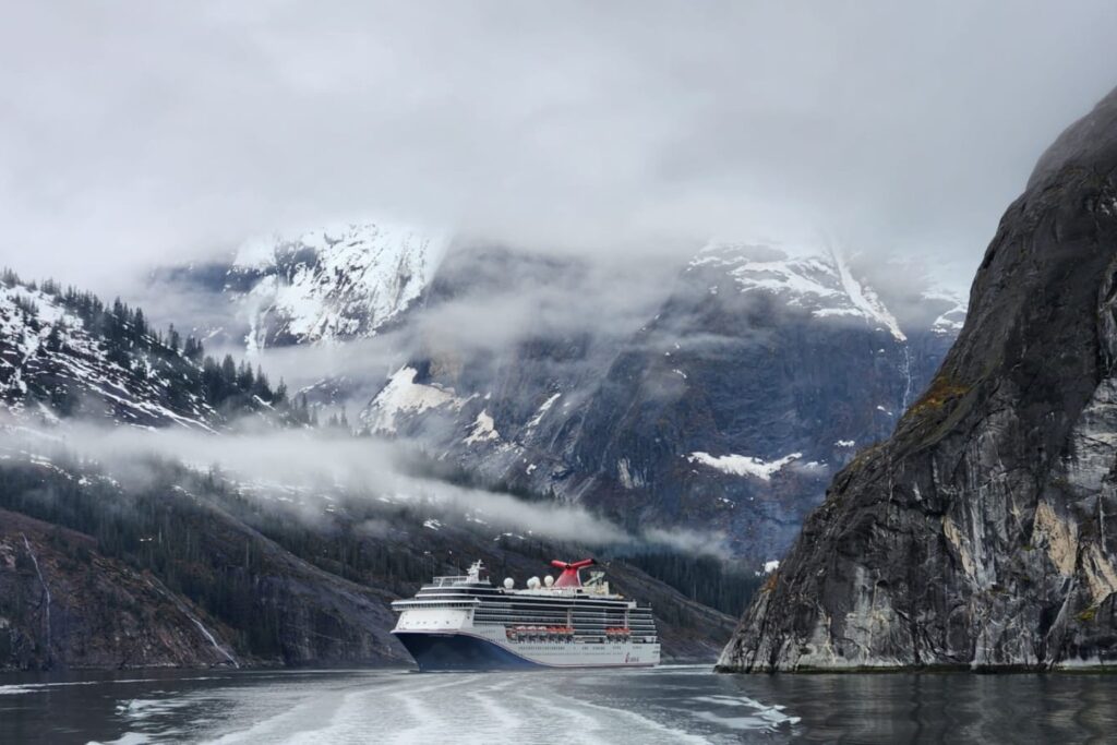 A Carnival Spirit cruise ship sails through a narrow Alaskan fjord, surrounded by misty snow-capped mountains and rocky cliffs.