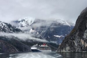 A Carnival Spirit cruise ship sails through a narrow Alaskan fjord, surrounded by misty snow-capped mountains and rocky cliffs.